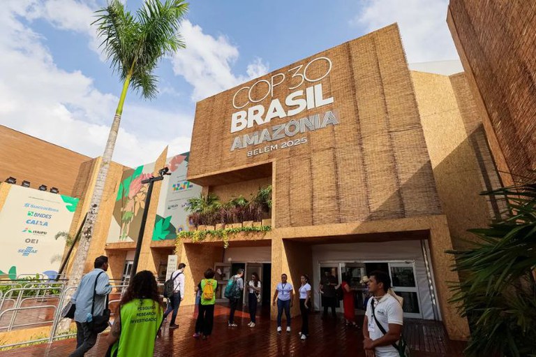 Fachada de um prédio coberto de madeira e bambu com os dizeres "COP30 Brasil Amazônia Belém 2025". Pessoas circulam na entrada.