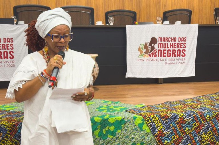 Mulher negra de roupa e turbante branco discursa ao microfone em evento com a faixa "Marcha das Mulheres Negras por Reparação e Bem Viver Brasília | 2025" e tecido estampado.