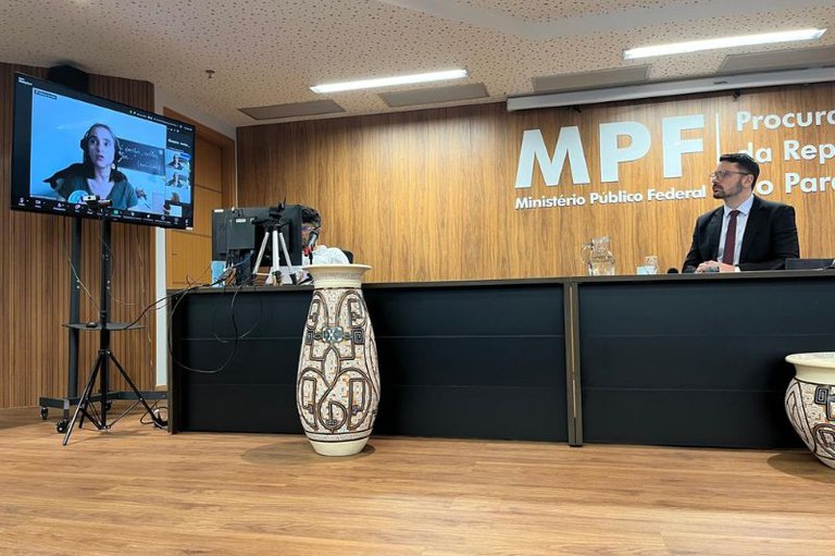 Foto do auditório do Ministério Público Federal (MPF) em Belém (PA). Há duas pessoas sentadas atrás de uma mesa preta, com um painel de madeira ao fundo exibindo a logo do MPF / Procuradoria da República no Pará. À esquerda, há uma tela com uma videoconferência ativa e dois vasos decorativos em frente à mesa.