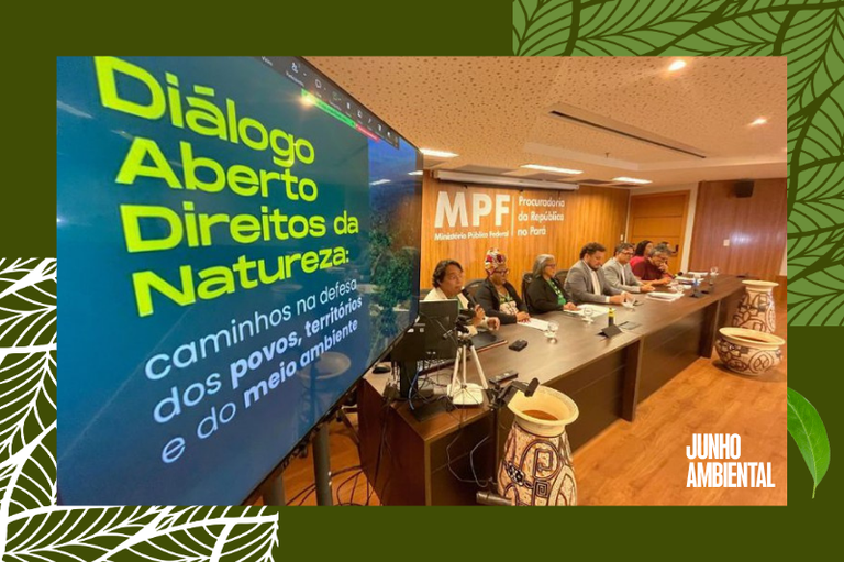 Foto em ângulo oblíquo mostra o painel de um evento em uma grande mesa com palestrantes e, em primeiro plano, uma tela de TV com o texto "Diálogo Aberto Direitos da Natureza: caminhos na defesa dos povos, territórios e do meio ambiente".