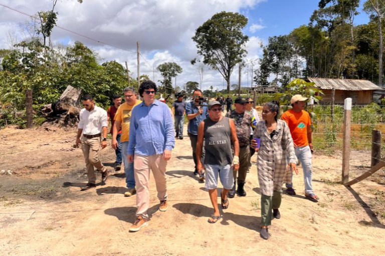 Um grupo de pessoas caminha por uma estrada de terra cercada por vegetação e algumas construções simples de madeira. Entre os presentes estão homens e mulheres com roupas casuais e uniformes, além de uma pessoa com câmera filmando. O céu está parcialmente nublado, com nuvens brancas e céu azul ao fundo. Há árvores altas e uma cerca de arame ao lado direito da imagem.