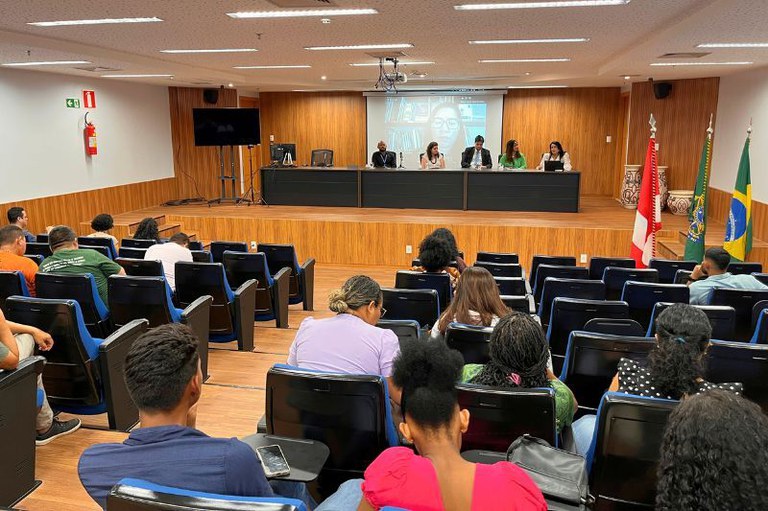 Foto de evento no auditório do MPF em Belém, que tem paredes de madeira clara. No palco, um painel de palestrantes está sentado em uma mesa, enquanto uma tela de projeção exibe outra participante por videochamada. O público, visto de costas, assiste à apresentação a partir das cadeiras da plateia. Ao lado do palco, há bandeiras hasteadas, incluindo a do Brasil.