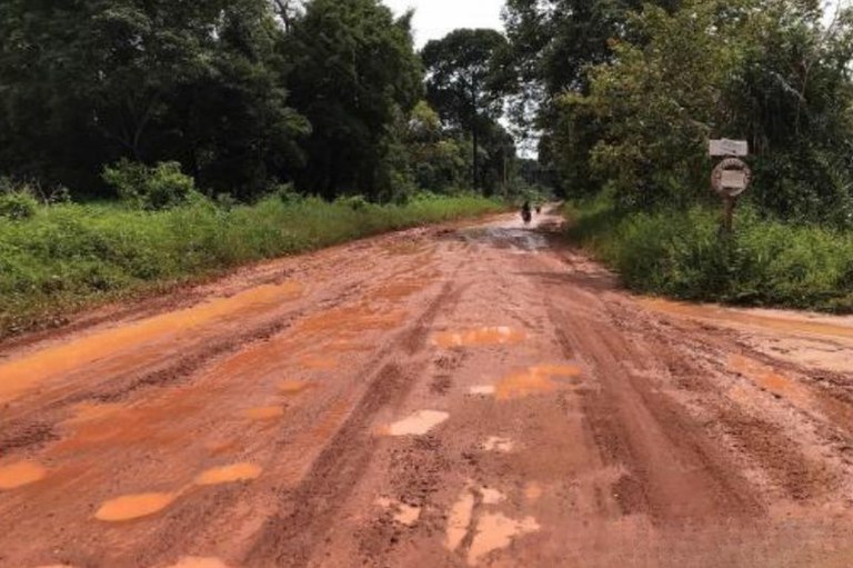 Foto de estrada de terra vermelha, lamacenta e esburacada, ladeada por vegetação densa (árvores e mato alto). Ao fundo, vê-se uma motocicleta e, à direita da via, uma placa de sinalização.