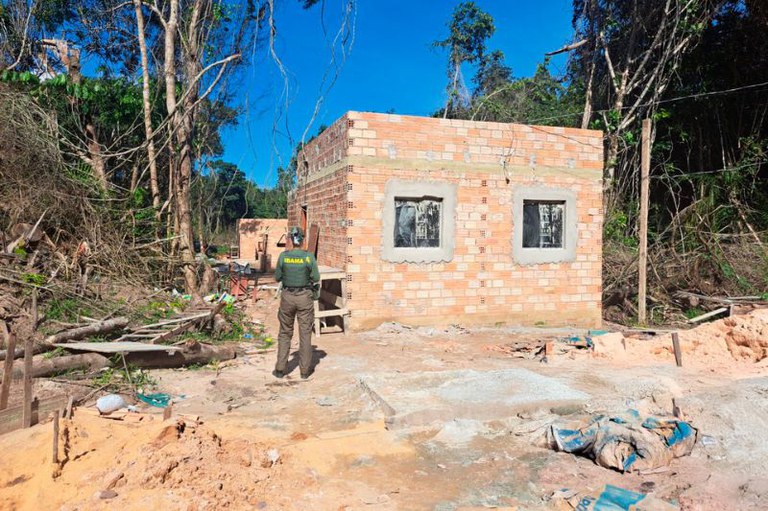 Foto em formato retangular. Um agente do Ibama, de costas, observa uma construção de tijolos aparentes, inacabada, com duas janelas na parede frontal. A cena se passa em uma área de mata, com árvores ao redor e indícios de desmatamento recente (troncos cortados e solo remexido). O céu está azul e limpo.