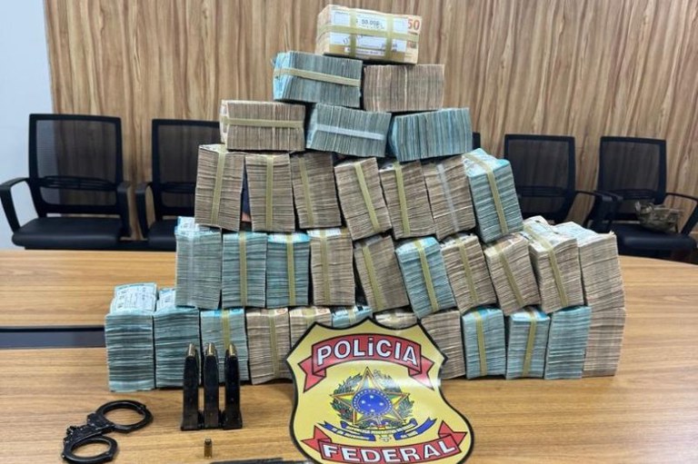 Pilhas de maços de dinheiro sobre mesa com algemas, carregadores de munição e um brasão da Polícia Federal ao centro.