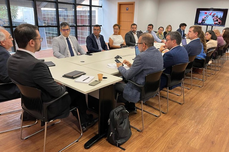 foto de um grupo de pessoas, predominantemente homens, vestidas com trajes formais, sentadas ao redor de uma grande mesa clara em uma sala de reuniões. Um homem em primeiro plano à esquerda está escrevendo em um caderno. Em frente a ele, outro homem manuseia um celular. Os demais participantes observam, e há notebooks e papéis sobre a mesa. Ao fundo, a sala possui grandes janelas e piso de madeira.