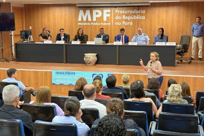 Pessoas em um auditório acompanham uma mulher de pé que fala ao microfone em frente a uma mesa com autoridades.