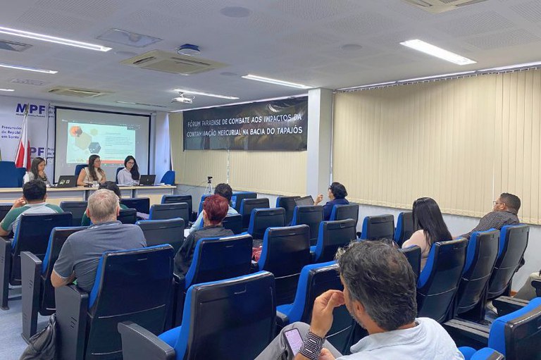 Pessoas sentadas em poltronas azuis do auditório do MPF em Santarém assistem a três mulheres em uma mesa de debate. Um banner atrás diz: "Fórum Paraense de Combate aos Impactos da Contaminação Mercurial na Bacia do Tapajós".