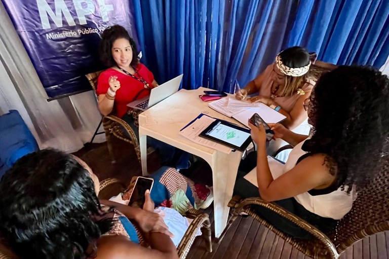 Foto vista de cima de quatro mulheres sentadas ao redor de uma mesa branca em um ambiente com cortinas azuis e um banner do MPF. Três estão trabalhando com notebooks, tablets e cadernos, e uma delas, de costas, olha para o celular.