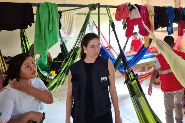 Foto do alojamento da Casa de Apoio à Saúde Indígena de Jacareacanga, espaço com várias redes penduradas em estruturas metálicas, roupas nas estruturas e pessoas presentes. Uma delas, a procuradora da República Thaís Medeiros da Costa, usa um colete azul com a logo do MPF.