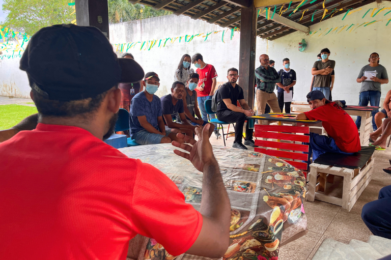 Foto mostra um grupo de pessoas reunidas sob uma cobertura rústica de madeira. No primeiro plano, de costas, um homem de camiseta vermelha e boné preto gesticula com a mão direita enquanto fala voltado para o grupo. Ao fundo, homens sentados em cadeiras azuis e outros de pé olham para ele