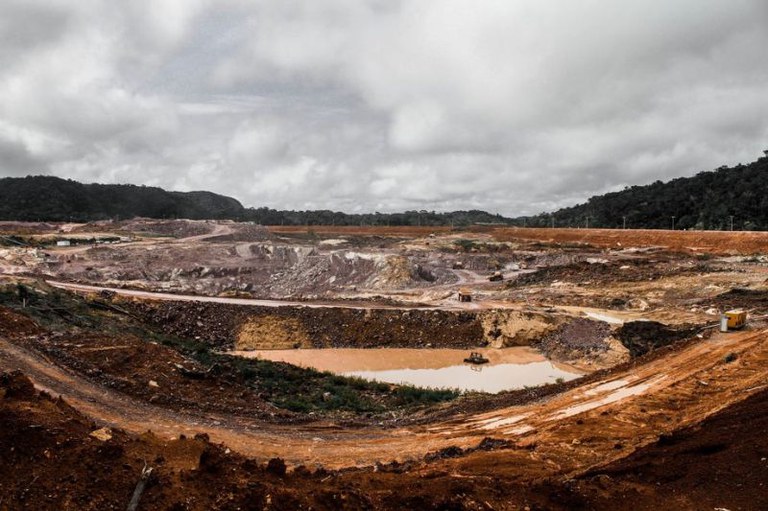 Foto de um terreno com terra em tons de laranja e marrom, com uma cratera cheia de lama e máquinas.