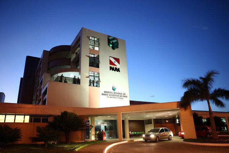 Foto da fachada do Hospital Regional do Baixo Amazonas, tirada à noite, com bandeira do Pará, luzes acesas, carros na entrada e céu azul escuro.
