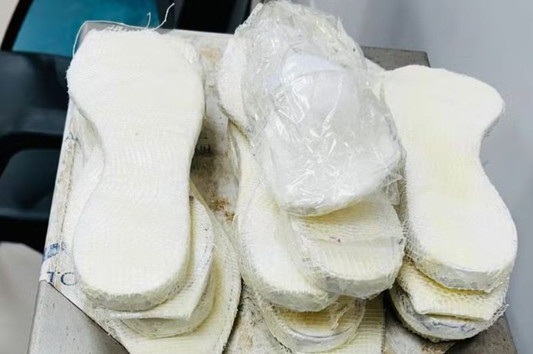 Foto mostra diversas palmilhas brancas e grossas empilhadas sobre uma superfície metálica, com um pacote plástico contendo cocaína acima delas.