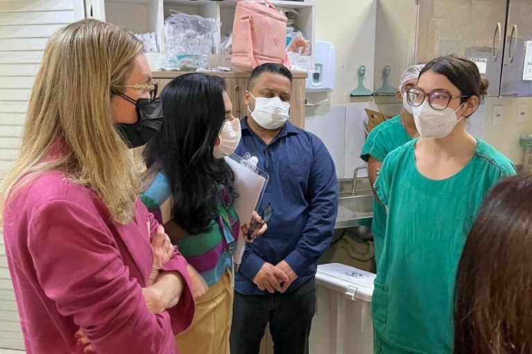 Pessoas com máscaras, jalecos verdes e roupas casuais conversam em uma sala com armários e materiais médicos ao fundo.