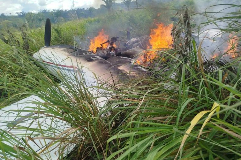 Foto de avião monomotor de cor clara caído em meio a um mato alto. A parte central da fuselagem está destruída, com fumaça e fogo.