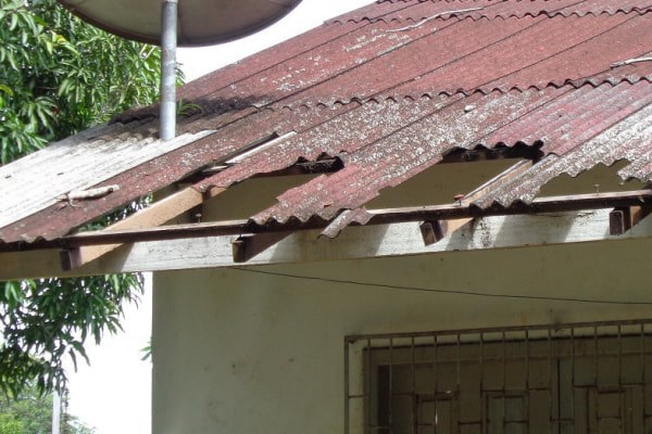 foto de detalhe de um telhado de amianto vermelho desgastado e quebrado, com partes faltando, sobre uma parede branca e uma janela gradeada.