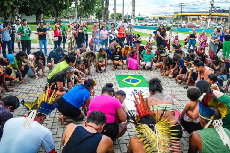 foto de pessoas, muitas delas indígenas com cocares, agachadas em círculo em volta de uma bandeira do Brasil no chão. Um público observa ao redor