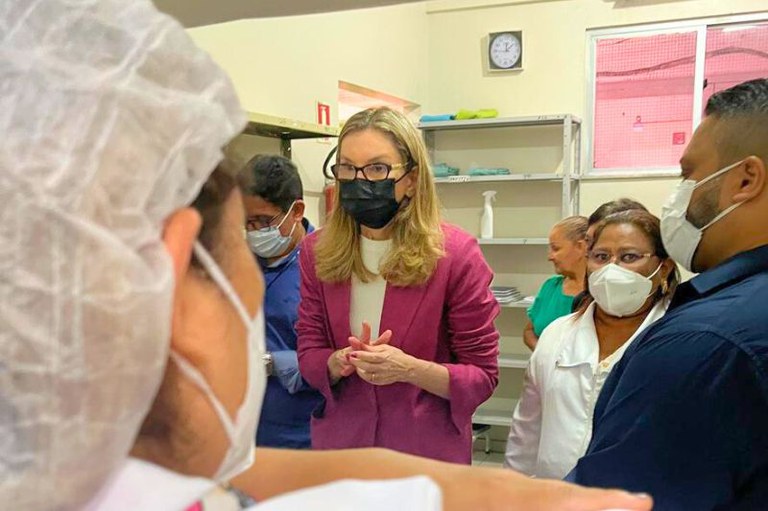 A foto mostra uma mulher loira de óculos, blazer magenta e máscara preta, conversa com grupo de pessoas de máscaras em sala com estantes