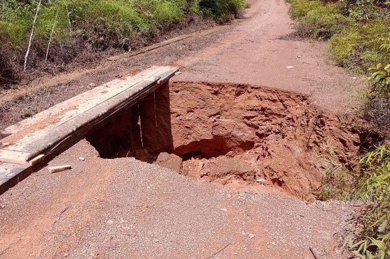 #DescriçãoDaImagem #Acessibilidade #ParaTodosVerem: Estrada de terra entre vegetação com um buraco profundo. Sobre o lado esquerdo da cratera, uma ponte de tábuas de madeira.