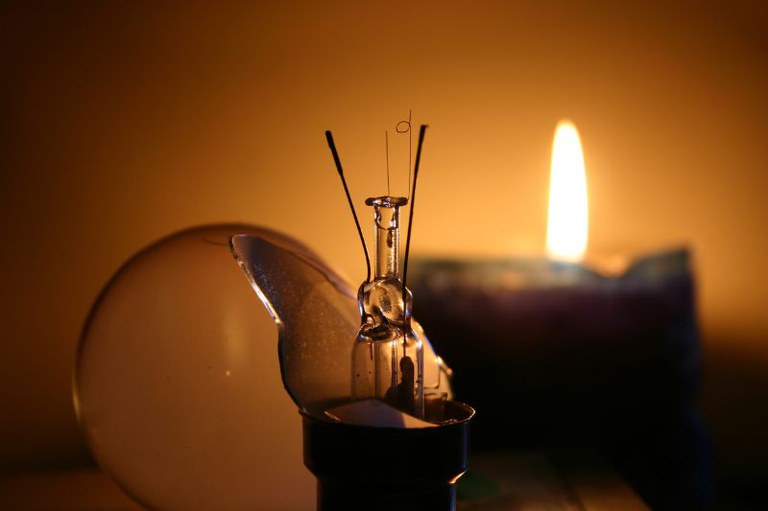 Foto mostra uma lâmpada quebrada com os filamentos expostos em primeiro plano, enquanto ao fundo há uma vela acesa.