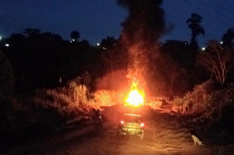 Foto noturna mostra carro em estrada de terra escura, com uma grande e intensa coluna de fogo no centro da cena.