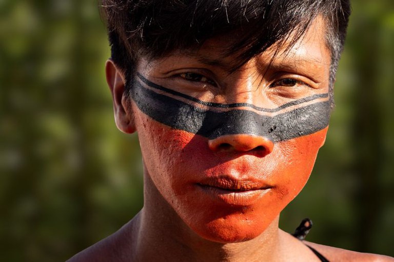 Jovem indígena com o rosto pintado de vermelho e preto, olhando para a câmera.