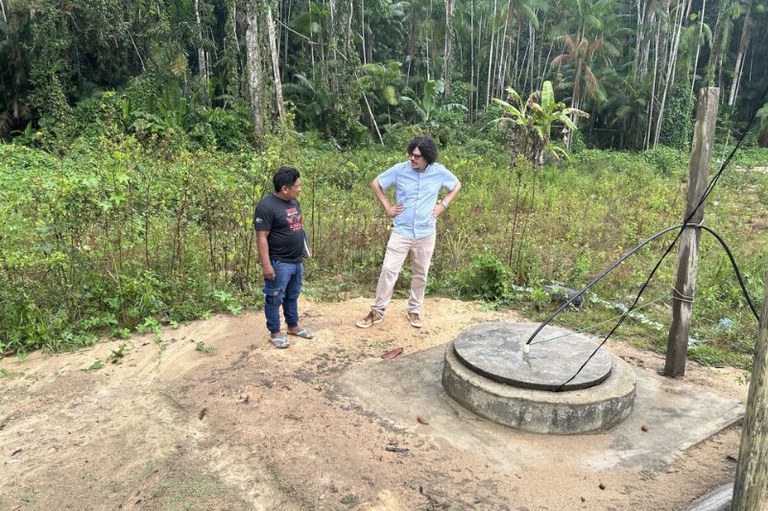 Dois homens, um indígena e um não indígena, conversam de pé em frente a um poço d'água no meio da floresta.
