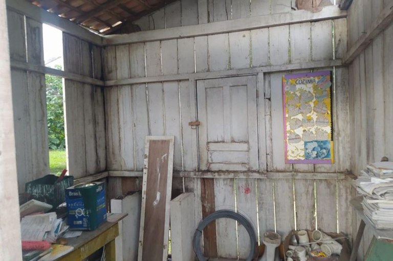 Interior de estrutura de escola, formada por madeira clara com uma pequena janela de madeira fechada, cartaz colorido, latas de tinta e objetos empilhados.