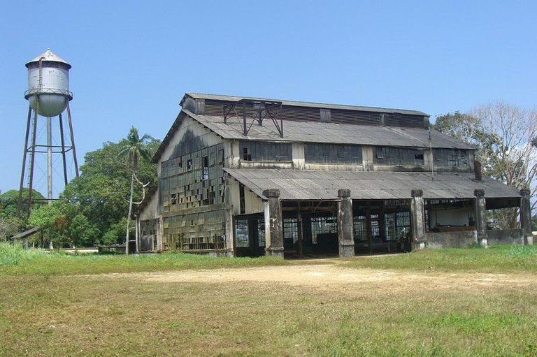 Galpão industrial abandonado com vidraças quebradas ao lado de uma alta caixa-d'água metálica em um terreno gramado sob céu azul em Fordlândia (PA)