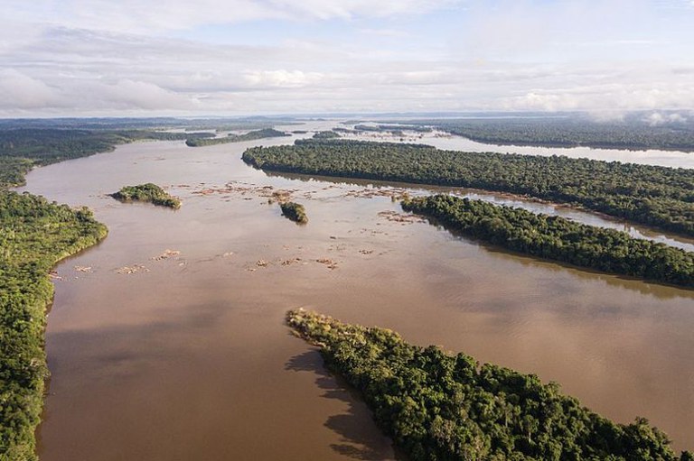 Foto aérea de rio largo de águas marrons, com ilhas e margens cobertas de mata verde densa, sob céu nublado.