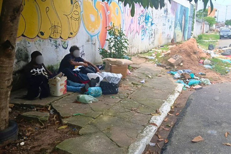 Duas pessoas sentadas na calçada de concreto junto a sacolas e latões, ao lado de muro com grafites e rua com lixo acumulado.