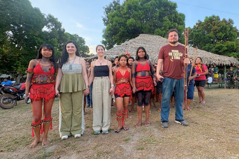 Seis pessoas em fila num chão de terra, entre elas indígenas de vestes vermelhas e um homem com camiseta Povos que escutam a terra.