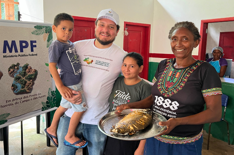 #ParaTodosVerem: Um homem de boné segura um menino no colo ao lado de duas mulheres, uma delas segurando uma grande bandeja de metal com um peixe assado, junto a um banner do Ministério Público Federal.