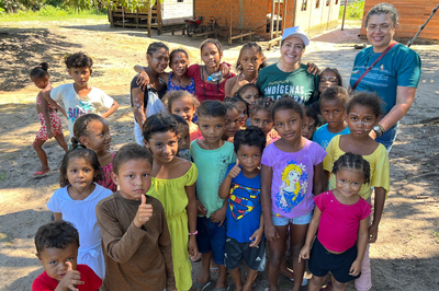 justica-itinerante-cooperativa-amazonia-legal-2026-2.png Grupo de dezenas de crianças indígenas e duas mulheres adultas posa sorrindo em um pátio de terra, à frente de casas simples de madeira e tijolos sob a luz do sol.