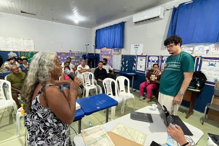 Mulher idosa com cabelos grisalhos fala ao microfone em uma sala de aula com público sentado ao fundo e um homem em pé ao lado de uma mesa com notebook acompanhando a apresentação.