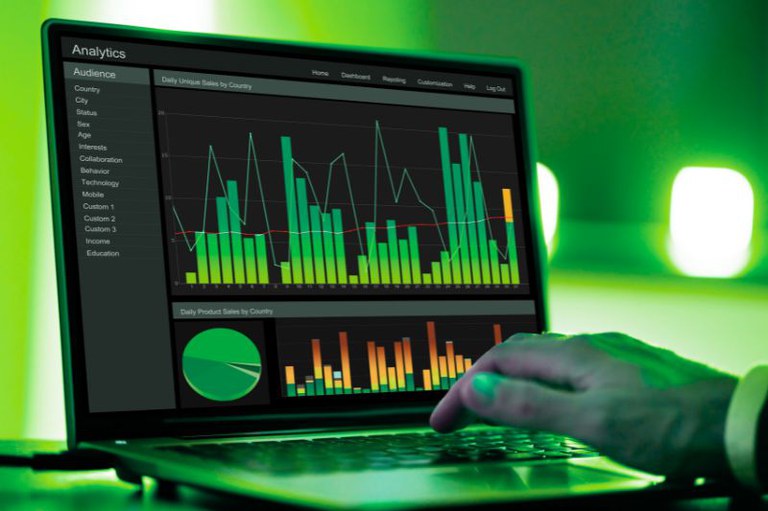 Foto em close-up da mão direita de uma pessoa digitando em um laptop que exibe gráficos e dados em uma tela escura e verde.