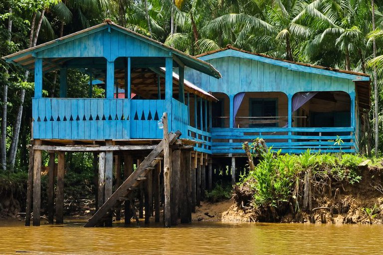 Casa de madeira azul construída sobre estacas de madeira à beira de um rio de água marrom, cercada por vegetação e palmeiras.