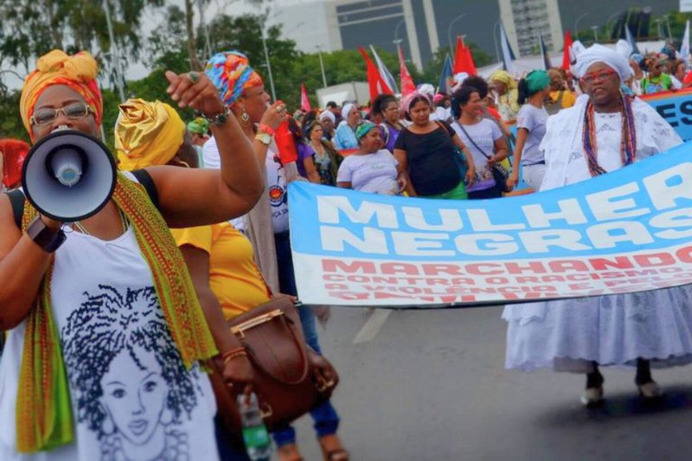 Foto colorida de uma marcha com mulheres negras. Uma mulher segura um megafone. Outras carregam uma faixa azul com texto em alusão à marcha.