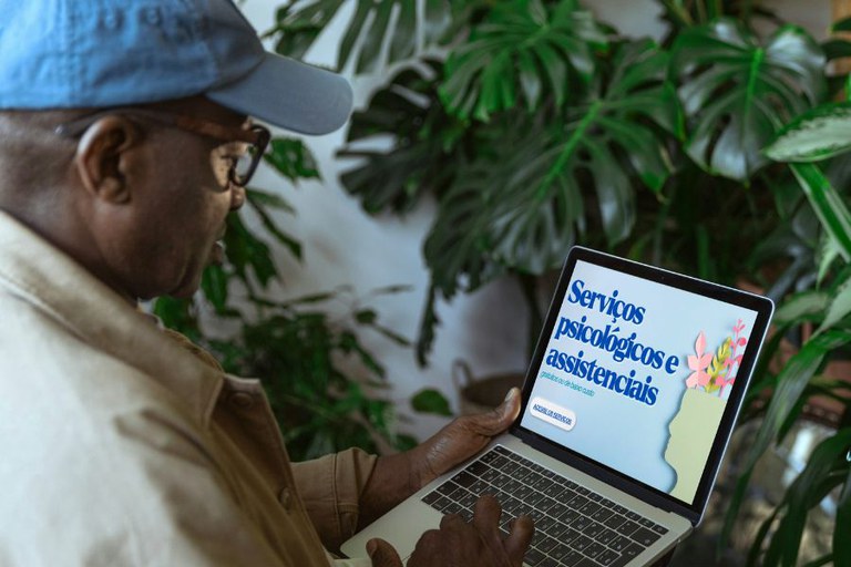 Foto de homem de boné azul e óculos que olha um notebook com o texto "Serviços psicológicos e assistenciais" em tela cercada por plantas.