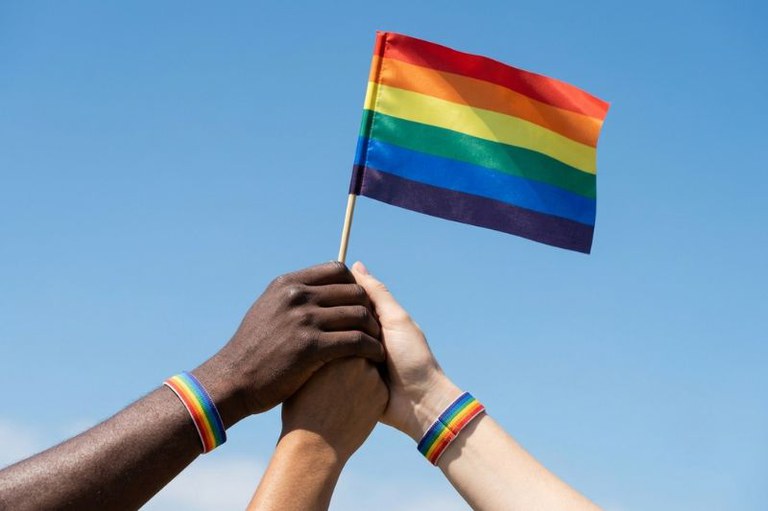 Três mãos de diferentes tons de pele erguidas seguram juntas uma pequena bandeira com as cores do arco-íris contra um céu azul.
