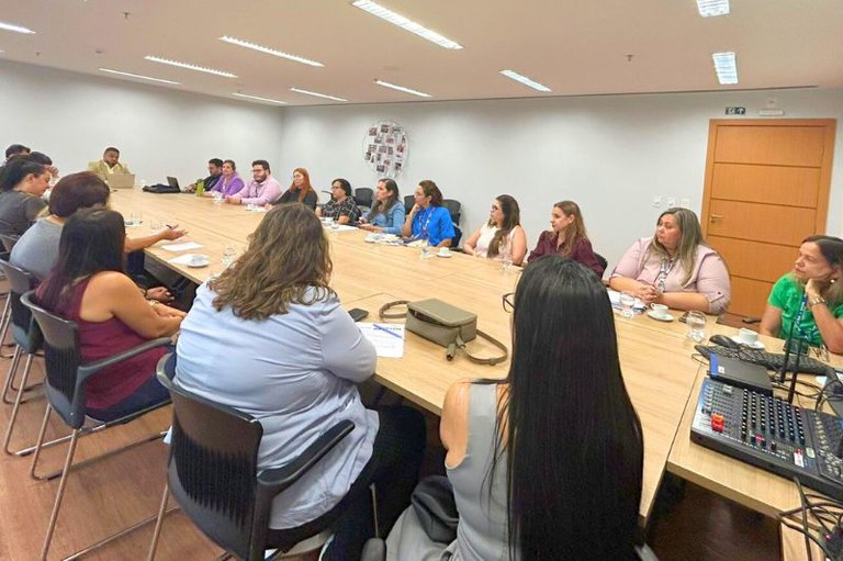 Reunião com cerca de vinte pessoas sentadas ao redor de uma grande mesa retangular em uma sala de reuniões iluminada, com paredes brancas e porta de madeira ao fundo.