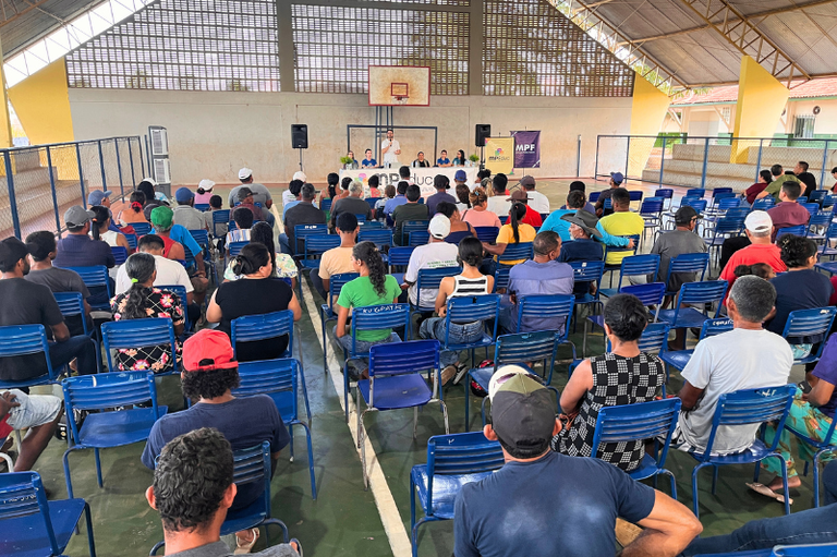 Vista traseira de pessoas sentadas em cadeiras azuis em uma quadra coberta, voltadas para uma mesa com autoridades e banners com os textos "MPEduc" e "MPF".