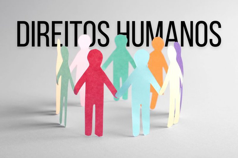 Foto de bonecos de papel coloridos em circulo, de mãos dadas, sob o texto "Direitos Humanos" em fundo cinza.