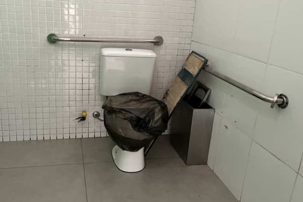Foto de um banheiro acessível fora de uso. O vaso sanitário está coberto por um saco de lixo preto. O ambiente possui paredes de azulejo branco, piso cinza e barras de apoio metálicas.