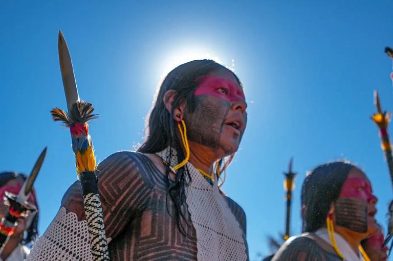 Uma pessoa indígena com o rosto e o corpo pintados em preto e vermelho olha para o lado, em um dia de sol forte. Ela segura uma lança decorada com penas coloridas, e ao fundo, outras pessoas e lanças são visíveis contra um céu azul intenso