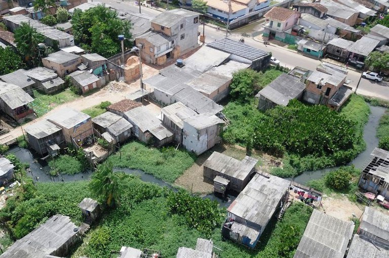 Fotografia aérea de casas de madeira e alvenaria às margens de um igarapé com vegetação em área urbana
