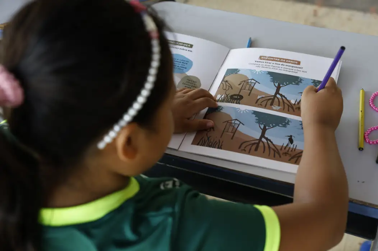 Vista superior traseira de uma criança de cabelos escuros e tiara de pérolas, sentada à mesa escolar. Ela observa e aponta para um livro didático aberto que apresenta uma atividade de "Encontre os Erros" sobre a preservação de manguezais. A criança segura um lápis roxo na mão direita.