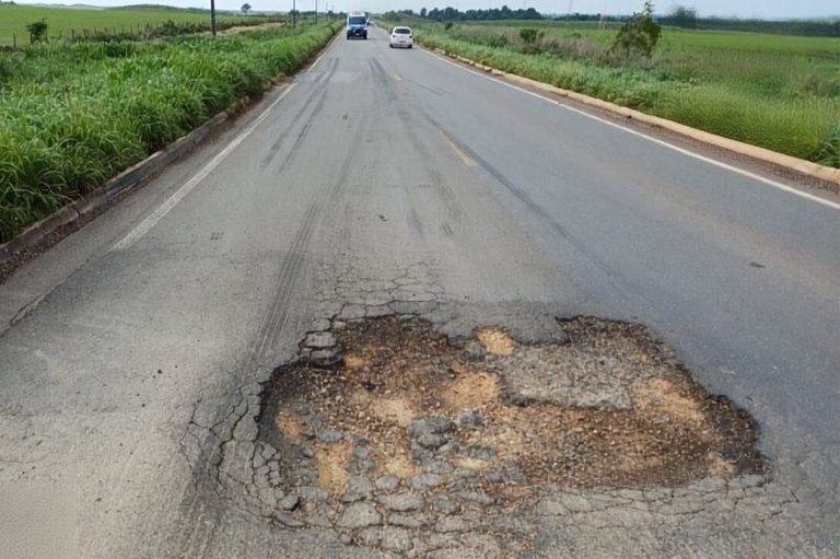 Foto em perspectiva de rodovia asfaltada com um grande buraco em primeiro plano revelando a terra por baixo. Ao fundo, dois veículos trafegam.
