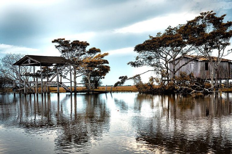 Casas de madeira sobre palafitas em um rio calmo, cercadas por árvores sob um céu nublado com reflexos na água.